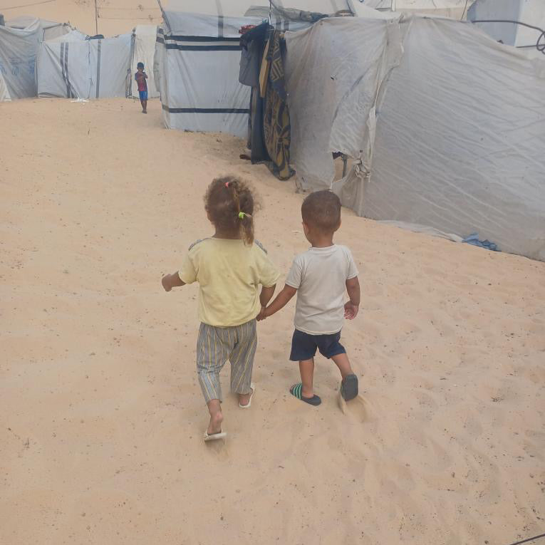 Two children holding hands on a sandy ground with tents in the background